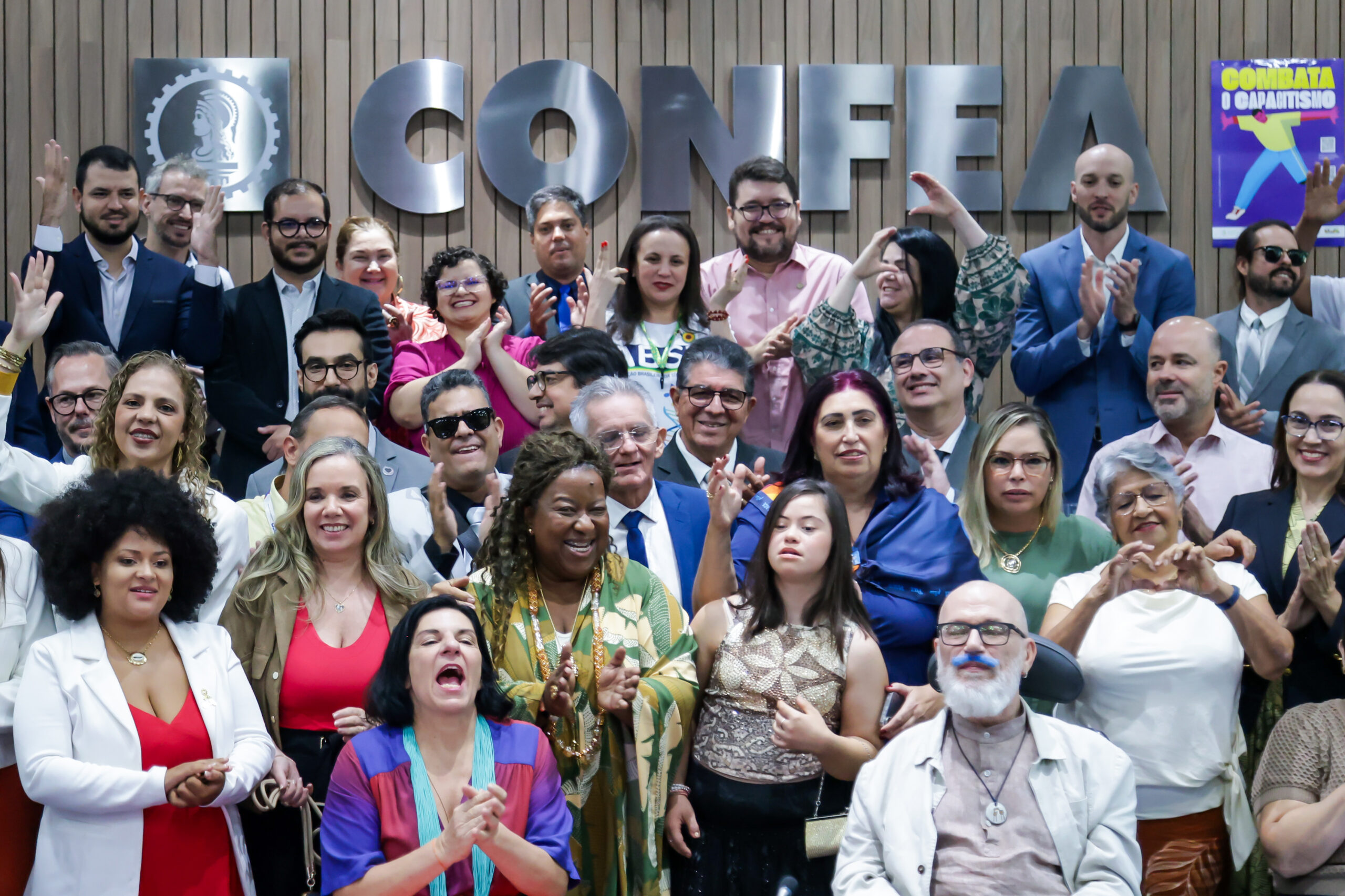 Conade empossa novos conselheiros Grupo diverso de pessoas sorrindo e comemorando em frente ao letreiro “CONFEA”. O ambiente transmite inclusão, com cartaz ao fundo escrito “Combata o capacitismo”.