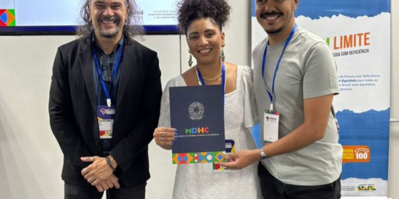 Três pessoas felizes, usando crachás, posam juntas em um evento institucional. A mulher ao centro segura uma pasta do Ministério dos Direitos Humanos e da Cidadania, em frente a banners do programa Viver Sem Limite.