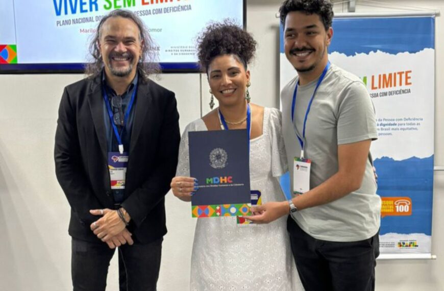 Três pessoas felizes, usando crachás, posam juntas em um evento institucional. A mulher ao centro segura uma pasta do Ministério dos Direitos Humanos e da Cidadania, em frente a banners do programa Viver Sem Limite.