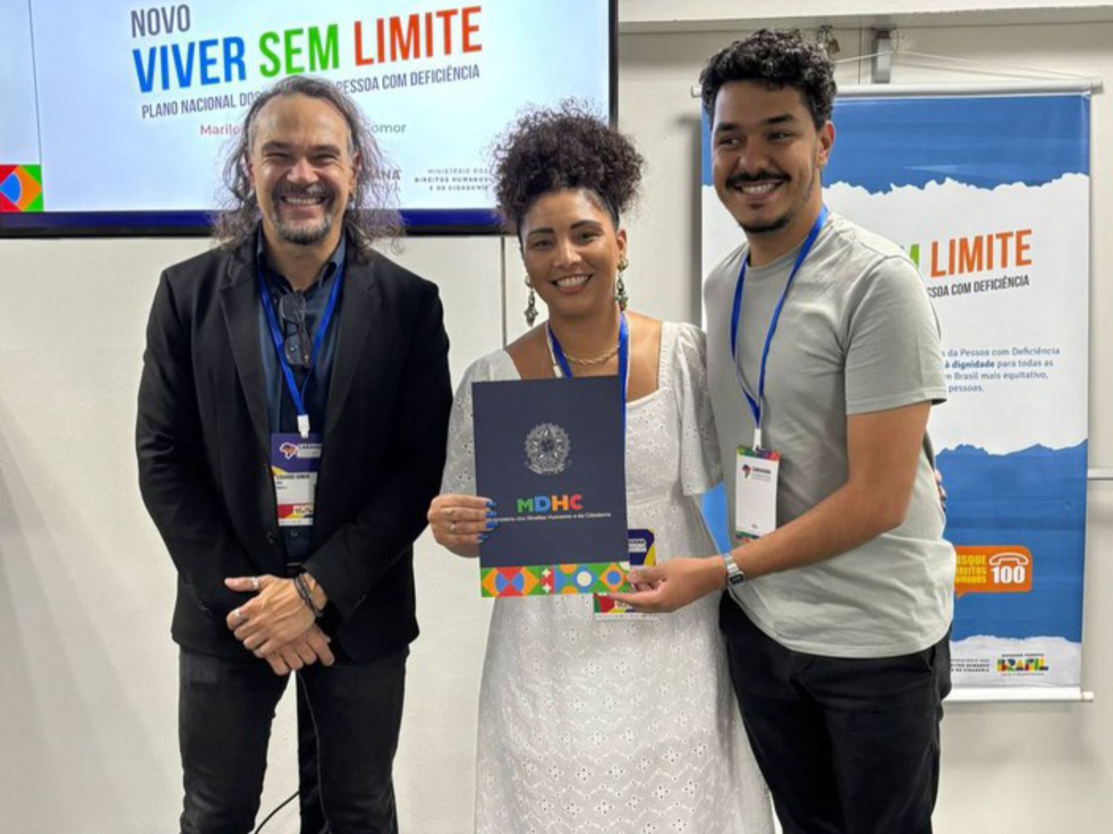 Três pessoas felizes, usando crachás, posam juntas em um evento institucional. A mulher ao centro segura uma pasta do Ministério dos Direitos Humanos e da Cidadania, em frente a banners do programa Viver Sem Limite.