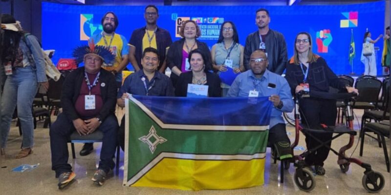 Grupo de pessoas posa em auditório de conferência, algumas sentadas e outras em pé, segurando uma bandeira. Há pessoas com deficiência, cadeiras acessíveis e palco iluminado ao fundo.