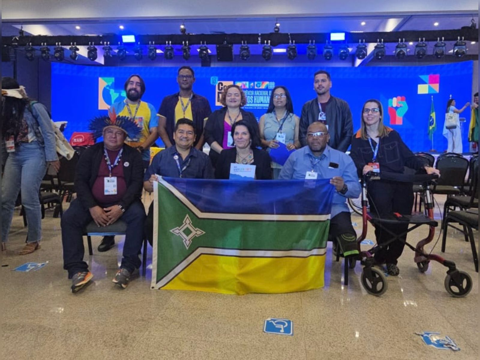Grupo de pessoas posa em auditório de conferência, algumas sentadas e outras em pé, segurando uma bandeira. Há pessoas com deficiência, cadeiras acessíveis e palco iluminado ao fundo.