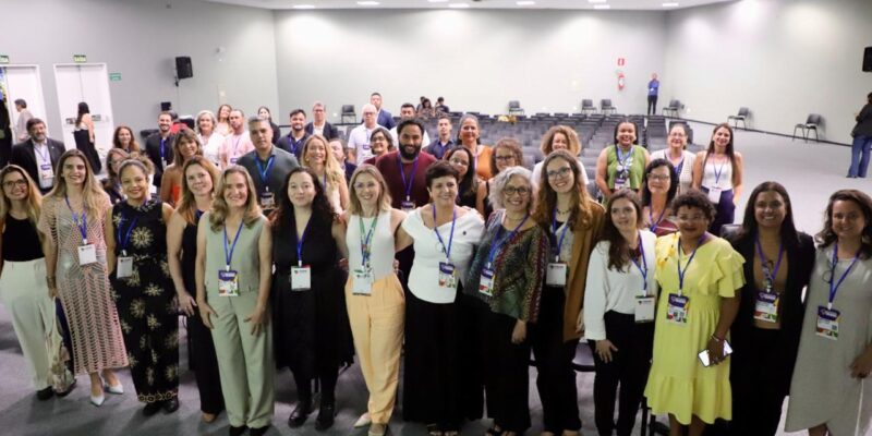 Uma grande equipe, composta por homens e mulheres com crachás no pescoço, está reunida em um salão iluminado. Todos estão sorrindo e olhando para a câmera, demonstrando união e profissionalismo.