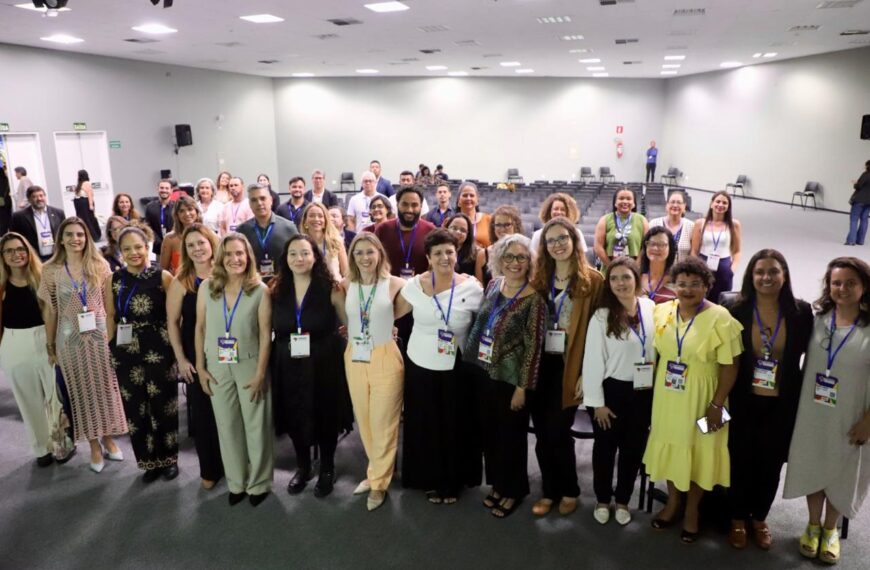 Uma grande equipe, composta por homens e mulheres com crachás no pescoço, está reunida em um salão iluminado. Todos estão sorrindo e olhando para a câmera, demonstrando união e profissionalismo.