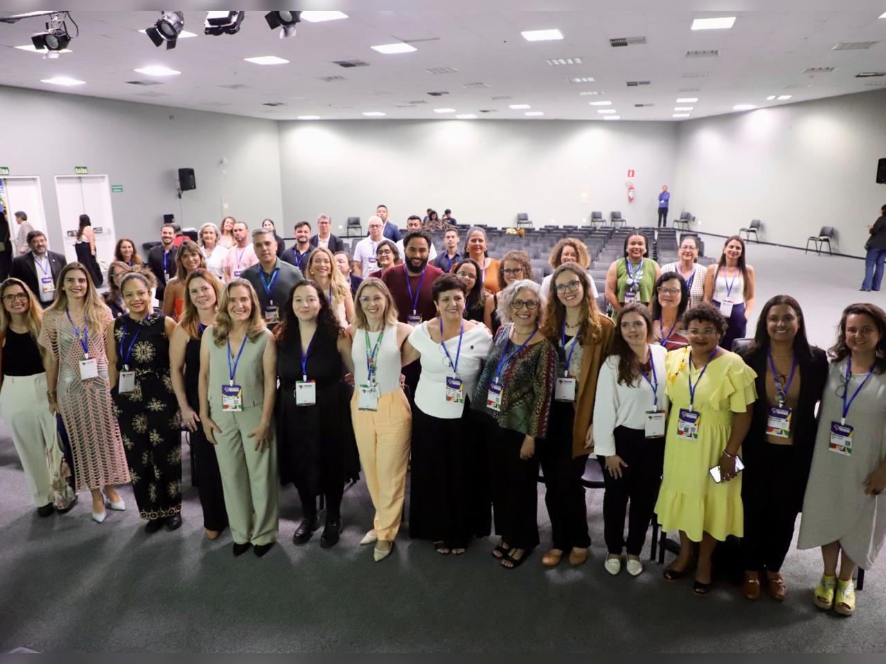 Uma grande equipe, composta por homens e mulheres com crachás no pescoço, está reunida em um salão iluminado. Todos estão sorrindo e olhando para a câmera, demonstrando união e profissionalismo.