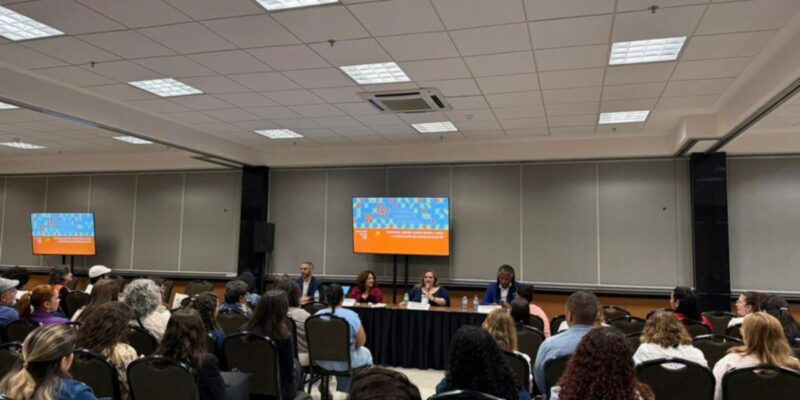 Auditório com várias pessoas assistindo a uma mesa-redonda composta por cinco palestrantes. Ao fundo, duas telas digitais mostram informações do evento. Ambiente organizado e iluminado.