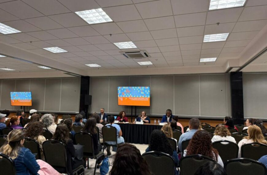Auditório com várias pessoas assistindo a uma mesa-redonda composta por cinco palestrantes. Ao fundo, duas telas digitais mostram informações do evento. Ambiente organizado e iluminado.