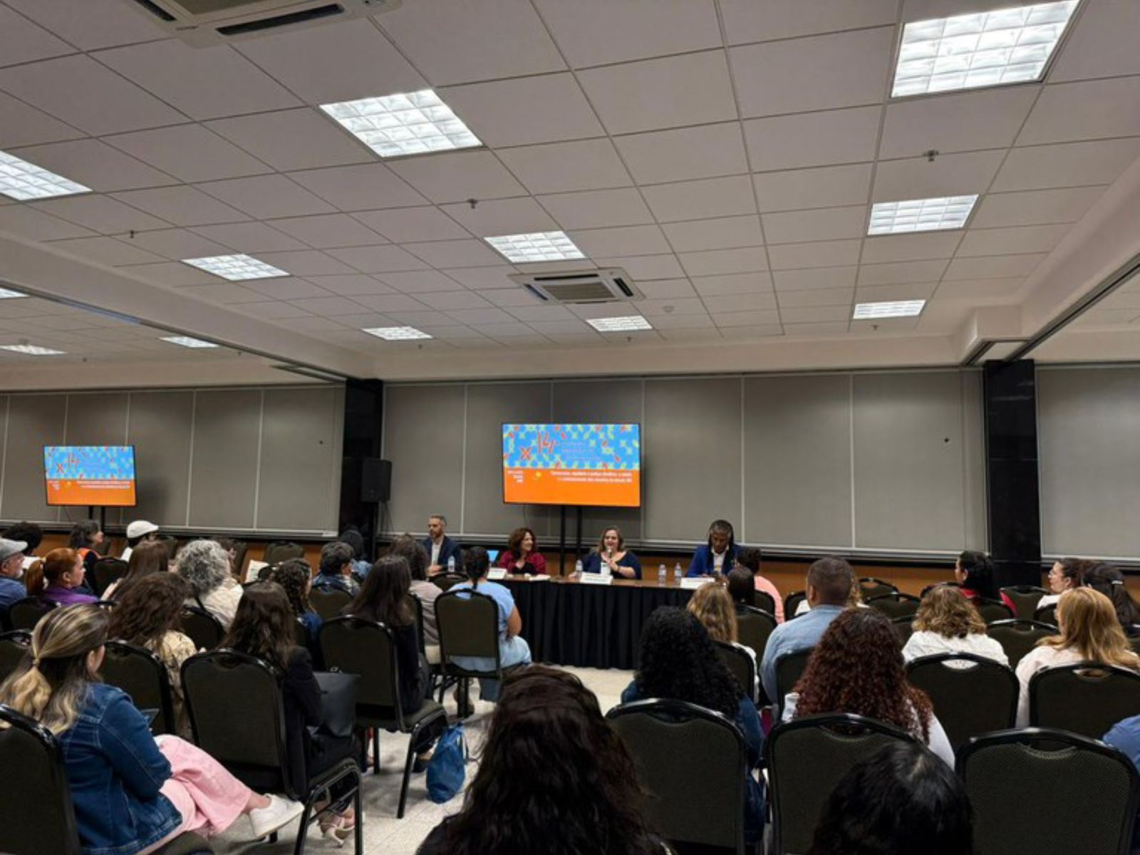 Auditório com várias pessoas assistindo a uma mesa-redonda composta por cinco palestrantes. Ao fundo, duas telas digitais mostram informações do evento. Ambiente organizado e iluminado.
