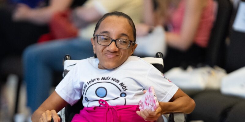 Mulher sorrindo em cadeira de rodas, usando óculos, camiseta branca com o texto "Nada Sobre Nós Sem Nós" e saia rosa. Ela segura um celular e há pessoas desfocadas sentadas ao fundo.