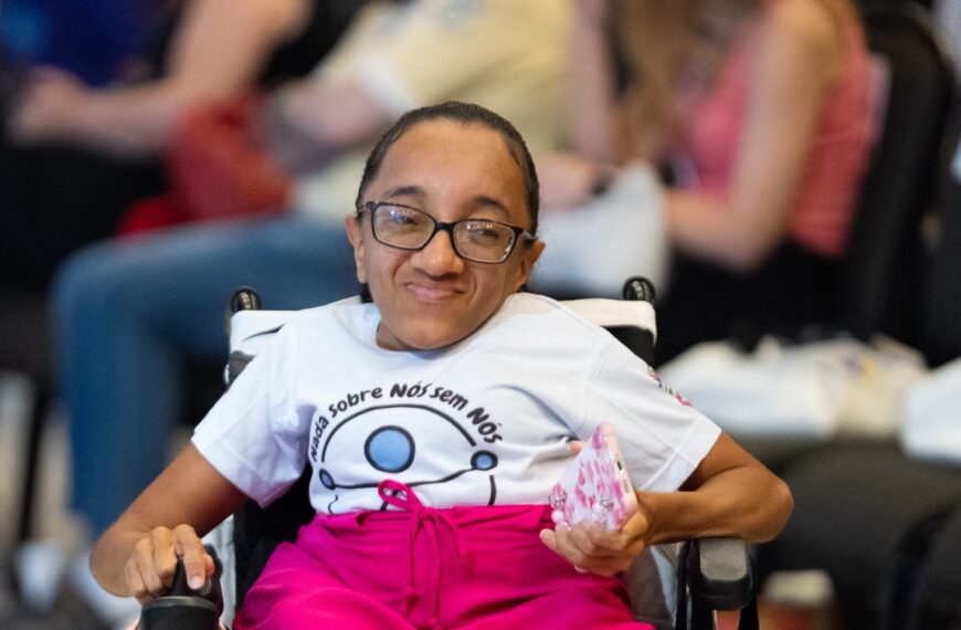 Mulher sorrindo em cadeira de rodas, usando óculos, camiseta branca com o texto "Nada Sobre Nós Sem Nós" e saia rosa. Ela segura um celular e há pessoas desfocadas sentadas ao fundo.