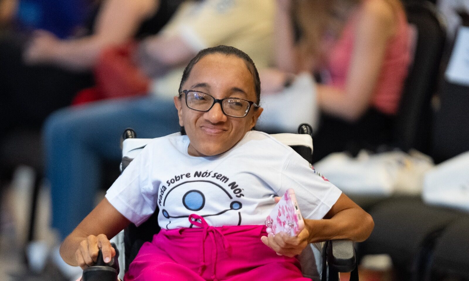 Mulher sorrindo em cadeira de rodas, usando óculos, camiseta branca com o texto "Nada Sobre Nós Sem Nós" e saia rosa. Ela segura um celular e há pessoas desfocadas sentadas ao fundo.