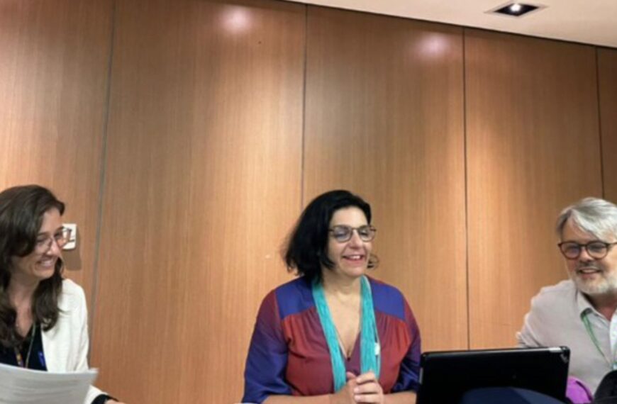 Três pessoas sentadas à mesa, sorrindo e conversando. Duas mulheres e um homem, todos de óculos, com papéis, notebook e garrafa de água, em uma sala com painel de madeira.