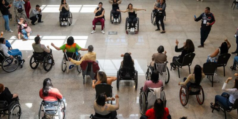 Várias pessoas, maioria em cadeiras de rodas, participam de uma atividade em círculo em um salão espaçoso e iluminado. Todas parecem envolvidas, algumas com os braços erguidos, em momento de integração.