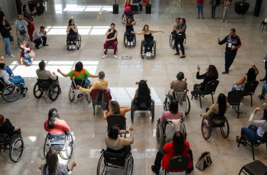Várias pessoas, maioria em cadeiras de rodas, participam de uma atividade em círculo em um salão espaçoso e iluminado. Todas parecem envolvidas, algumas com os braços erguidos, em momento de integração.