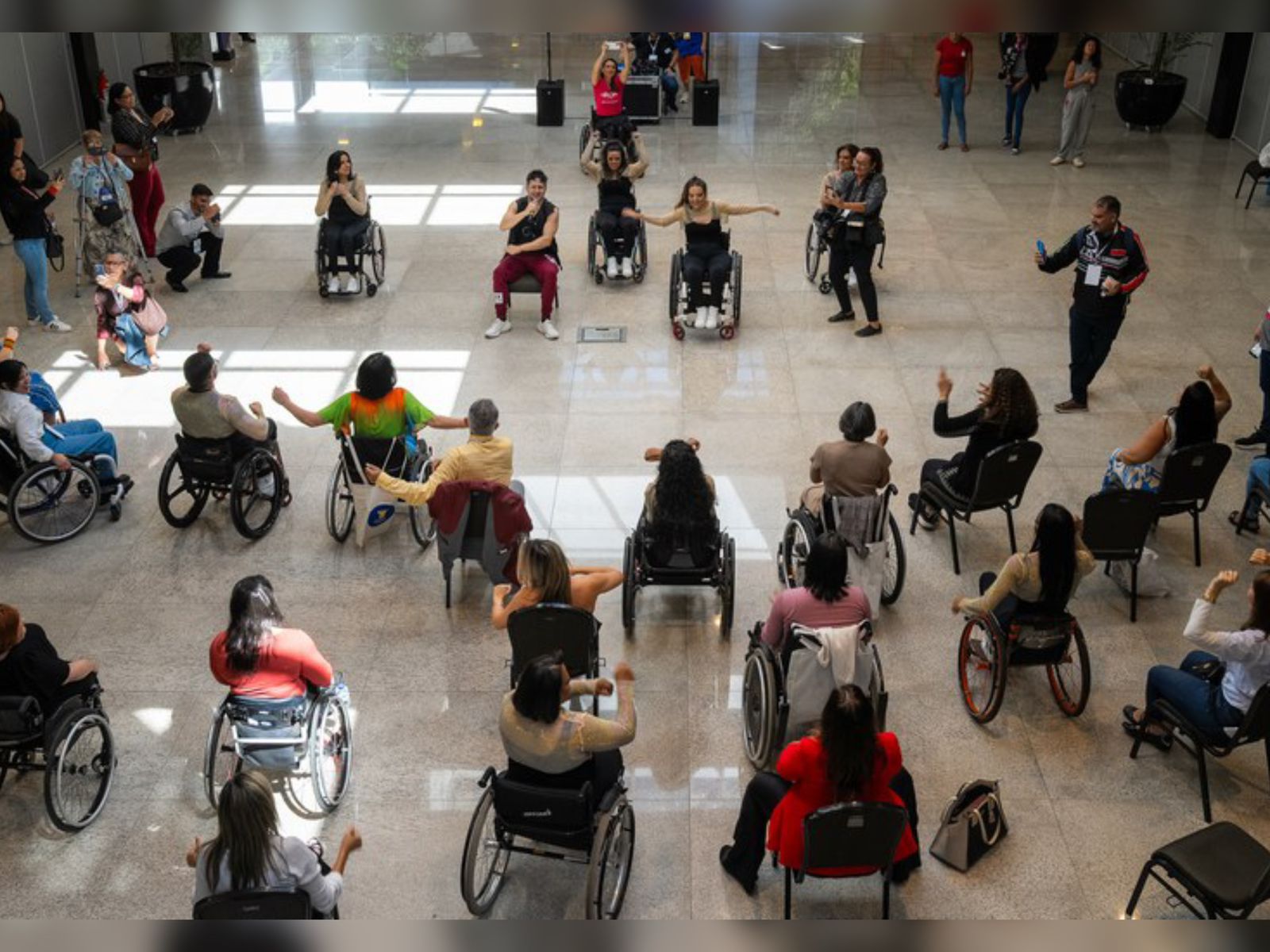 Várias pessoas, maioria em cadeiras de rodas, participam de uma atividade em círculo em um salão espaçoso e iluminado. Todas parecem envolvidas, algumas com os braços erguidos, em momento de integração.