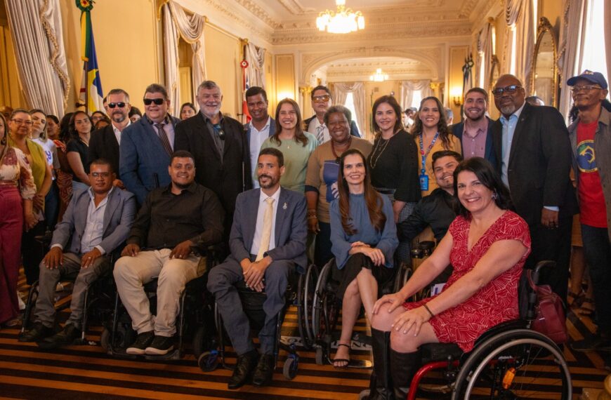 Foto de grupo em um salão elegante: pessoas com e sem deficiência, algumas em cadeiras de rodas, todas sorrindo. O ambiente é formal, com lustres, cortinas tradicionais e bandeiras ao fundo.
