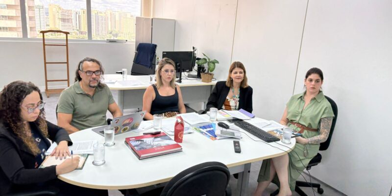 Cinco pessoas em reunião de trabalho em sala clara, com mesa oval, papéis, notebooks e copos d’água.