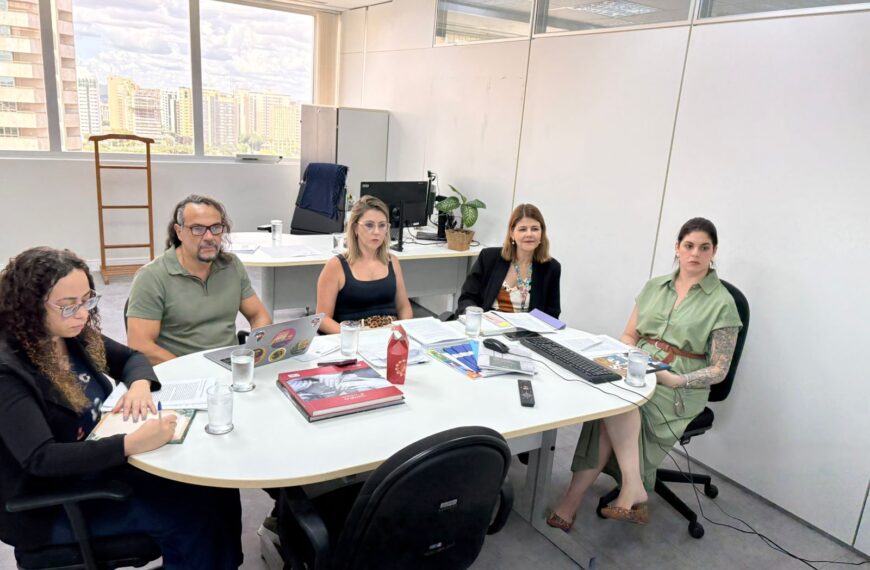 Cinco pessoas em reunião de trabalho em sala clara, com mesa oval, papéis, notebooks e copos d’água.