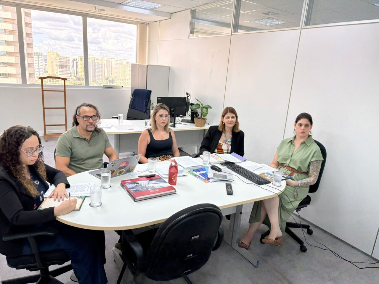Cinco pessoas em reunião de trabalho em sala clara, com mesa oval, papéis, notebooks e copos d’água.