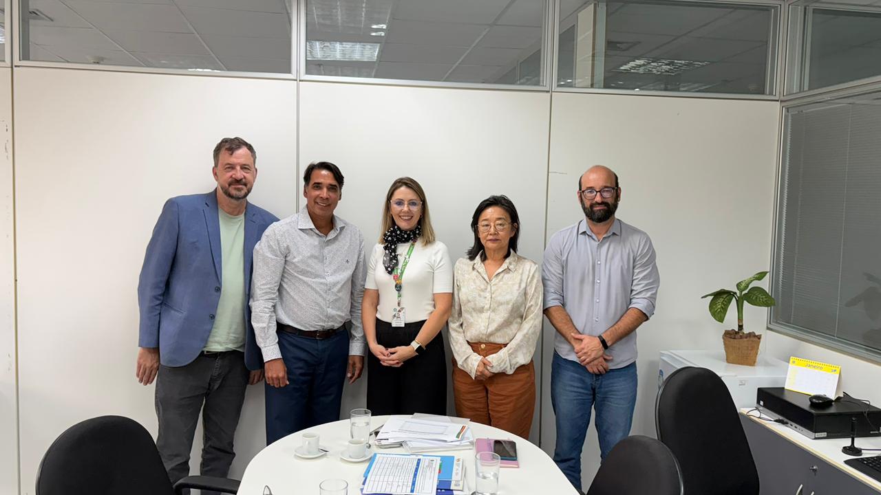 Cinco pessoas posam num escritório atrás de mesa com copos, xícaras, papéis; há planta ao lado.