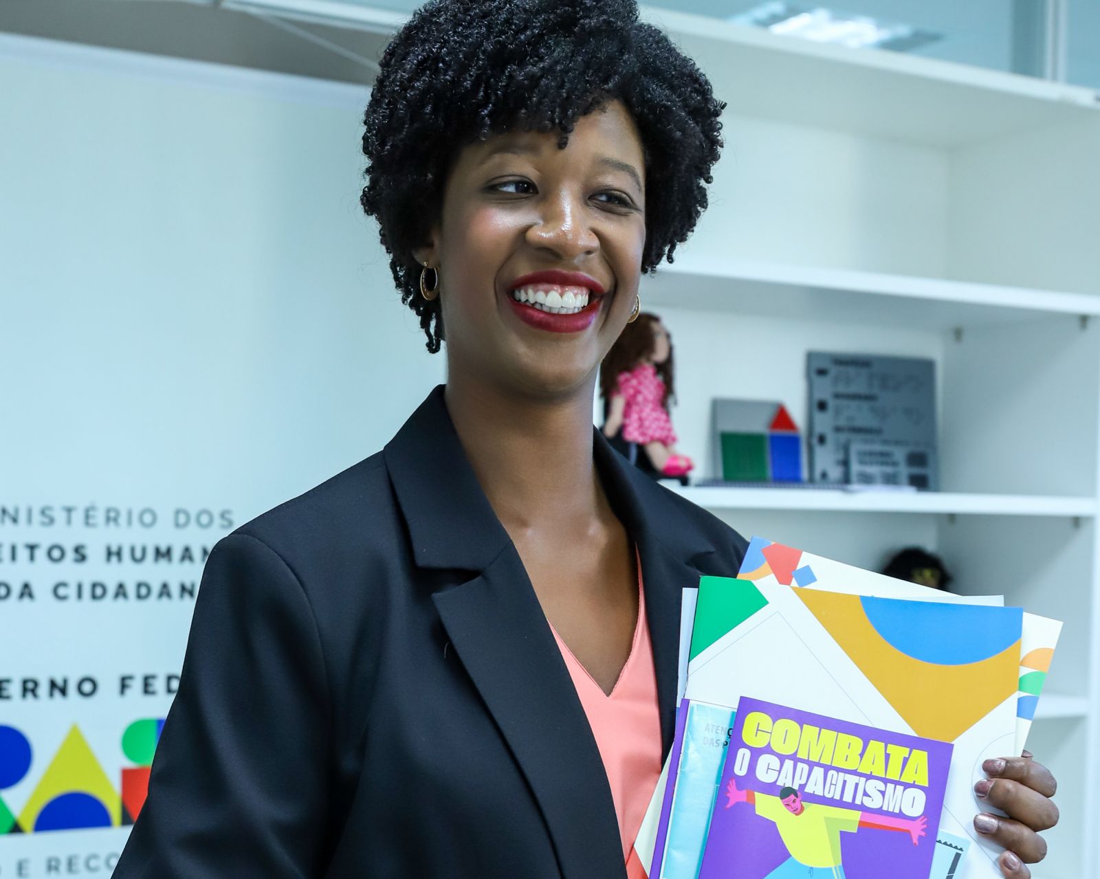 Histórias de vida e pertencimento: pessoas trans com deficiência fortalecem a mobilização por direitos no Brasil Mulher negra sorridente de blazer preto segura pasta colorida “Combate à Corrupção” em escritório.