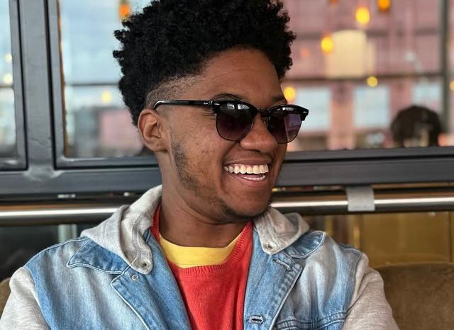 Histórias de vida e pertencimento: pessoas trans com deficiência fortalecem a mobilização por direitos no Brasil Homem rindo de óculos escuros, cabelo alto, jaqueta jeans com capuz; fundo de café/vidraça.