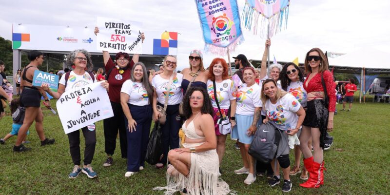 Grupo em evento ao ar livre com cartazes arco-íris; pessoa sentada à frente.