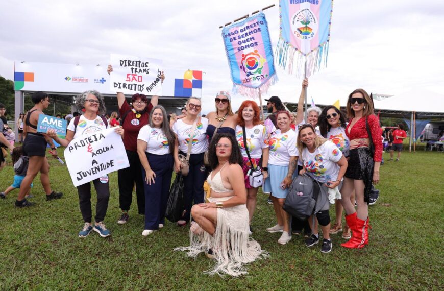 Grupo em evento ao ar livre com cartazes arco-íris; pessoa sentada à frente.