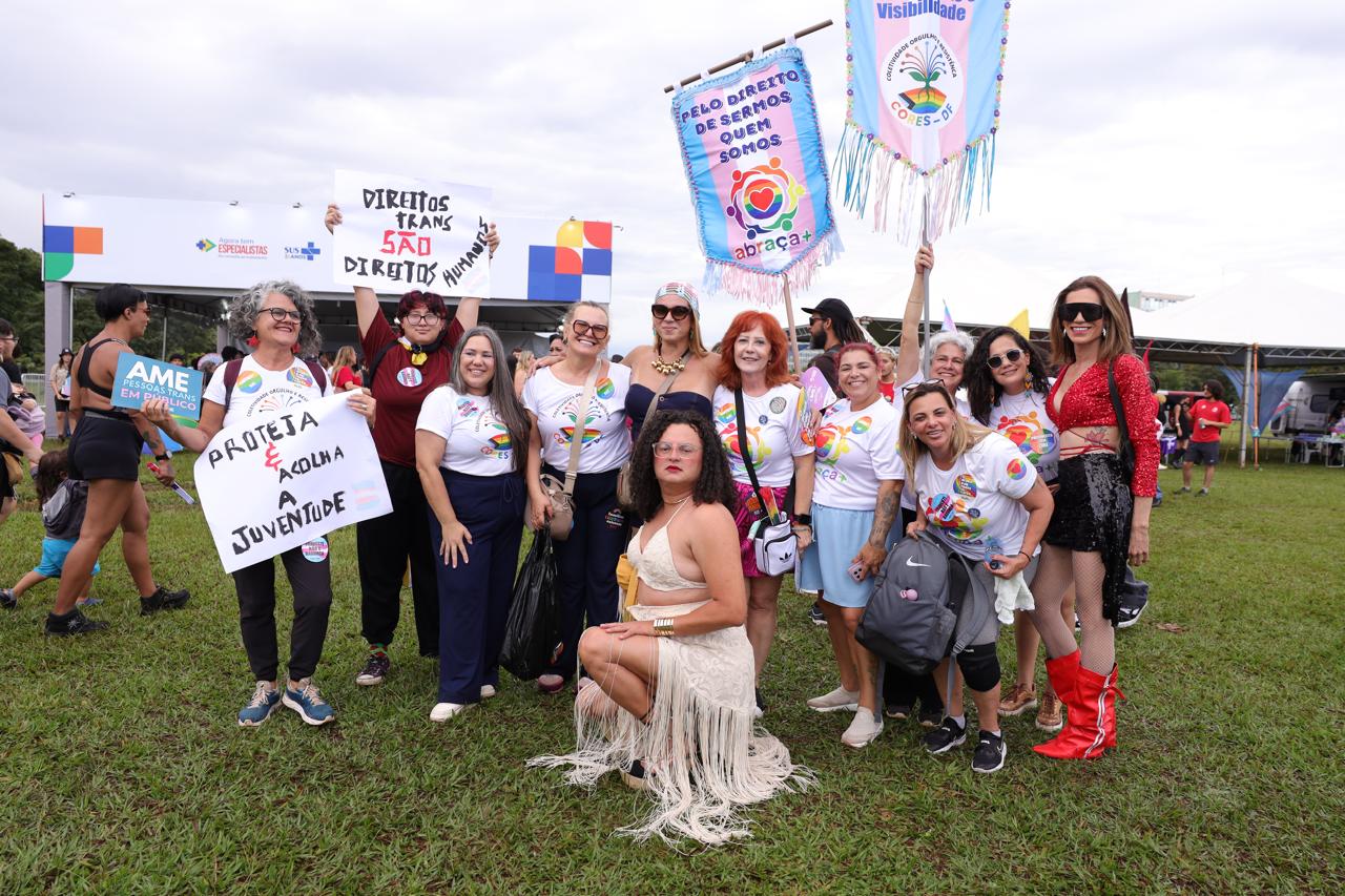 Grupo em evento ao ar livre com cartazes arco-íris; pessoa sentada à frente.
