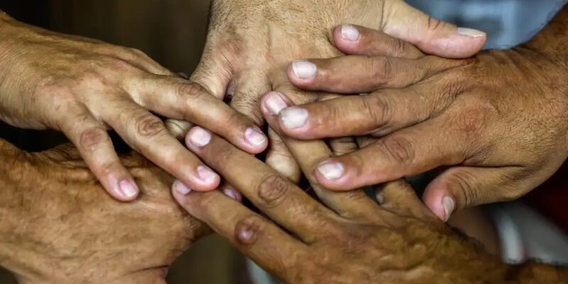 Cinco mãos de pessoas diferentes estão empilhadas no centro da foto, com dedos sobrepostos, sugerindo união e cooperação. A imagem é um close nas mãos; o fundo aparece desfocado, com roupas claras.