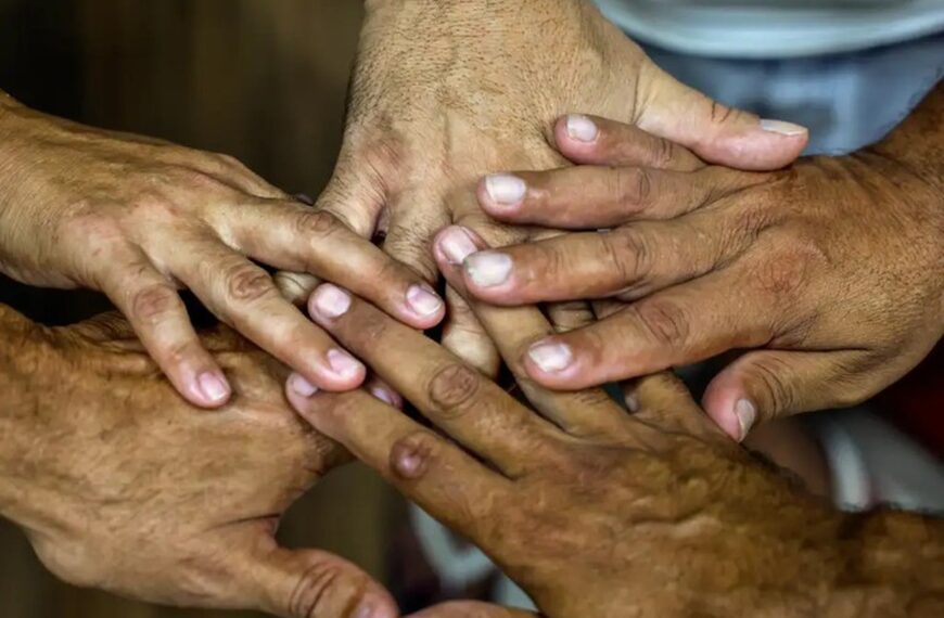 Cinco mãos de pessoas diferentes estão empilhadas no centro da foto, com dedos sobrepostos, sugerindo união e cooperação. A imagem é um close nas mãos; o fundo aparece desfocado, com roupas claras.
