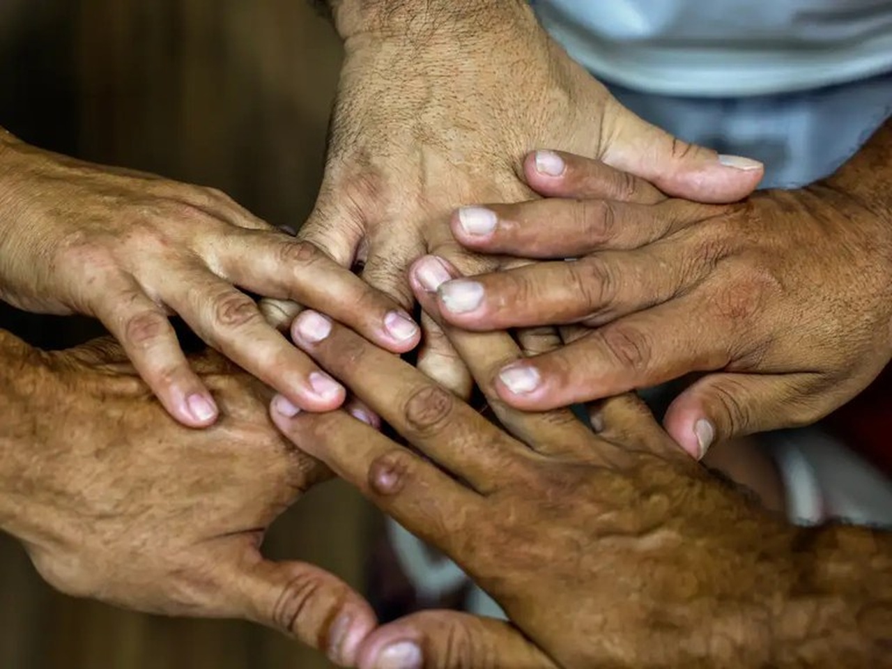 Cinco mãos de pessoas diferentes estão empilhadas no centro da foto, com dedos sobrepostos, sugerindo união e cooperação. A imagem é um close nas mãos; o fundo aparece desfocado, com roupas claras.