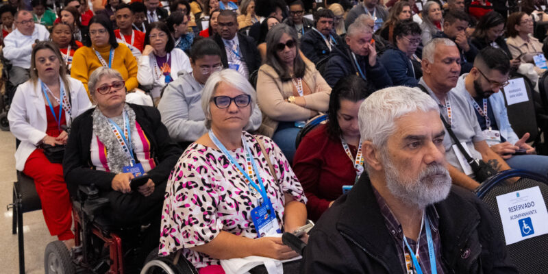 Plano frontal de auditório com público numeroso sentado em fileiras. Em primeiro plano, pessoas adultas, entre elas usuários de cadeira de rodas, utilizam crachás e observam a atividade com atenção. O espaço é amplo, iluminado e acessível.