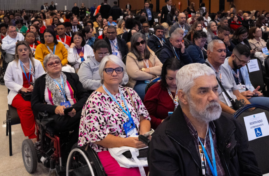 Plano frontal de auditório com público numeroso sentado em fileiras. Em primeiro plano, pessoas adultas, entre elas usuários de cadeira de rodas, utilizam crachás e observam a atividade com atenção. O espaço é amplo, iluminado e acessível.