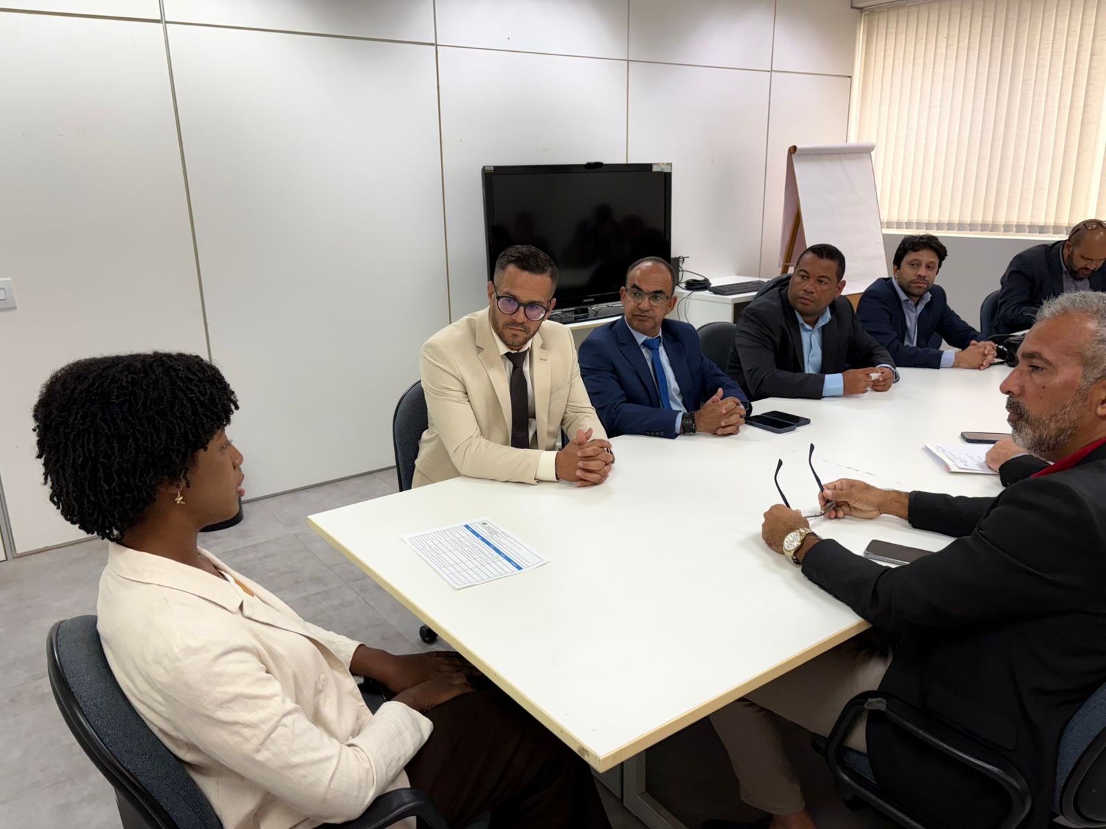 Sete pessoas participam de reunião em sala administrativa, sentadas ao redor de mesa de trabalho. Ambiente institucional com paredes claras, monitor e quadro de apoio ao fundo e iluminação artificial.