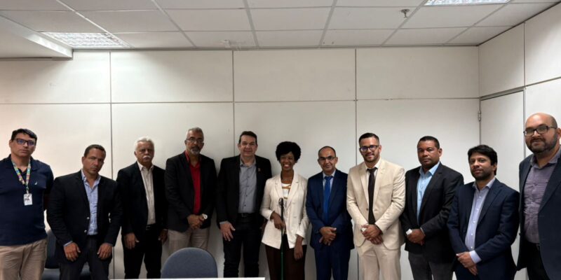 Reunião institucional em sala de escritório reúne onze pessoas em trajes formais, posicionadas atrás de mesa. Ao centro, mulher segura bengala. Ambiente administrativo com paredes claras e iluminação artificial.