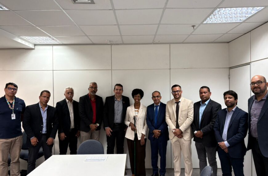 Reunião institucional em sala de escritório reúne onze pessoas em trajes formais, posicionadas atrás de mesa. Ao centro, mulher segura bengala. Ambiente administrativo com paredes claras e iluminação artificial.