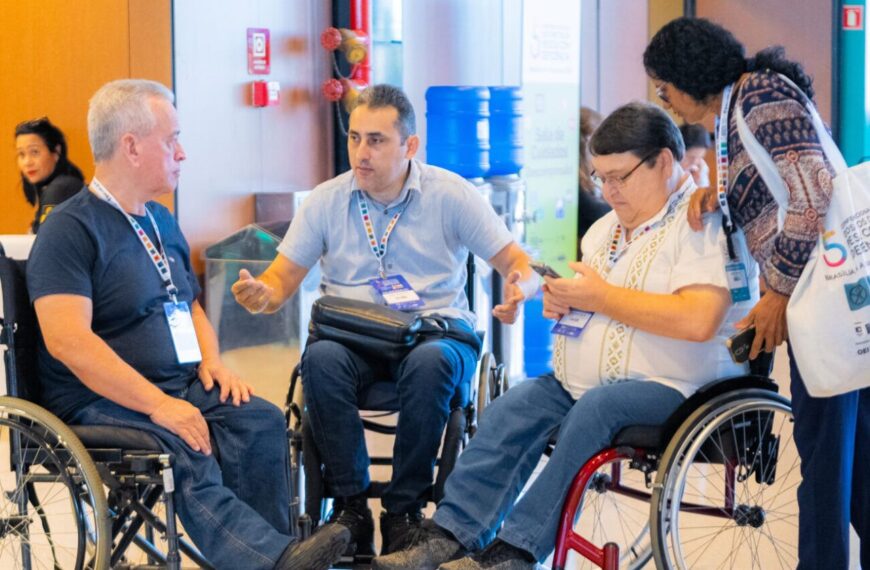 Três homens em cadeiras de rodas conversam em espaço interno de evento, enquanto uma mulher em pé interage com o grupo. Todos usam crachás e participam de atividade institucional.