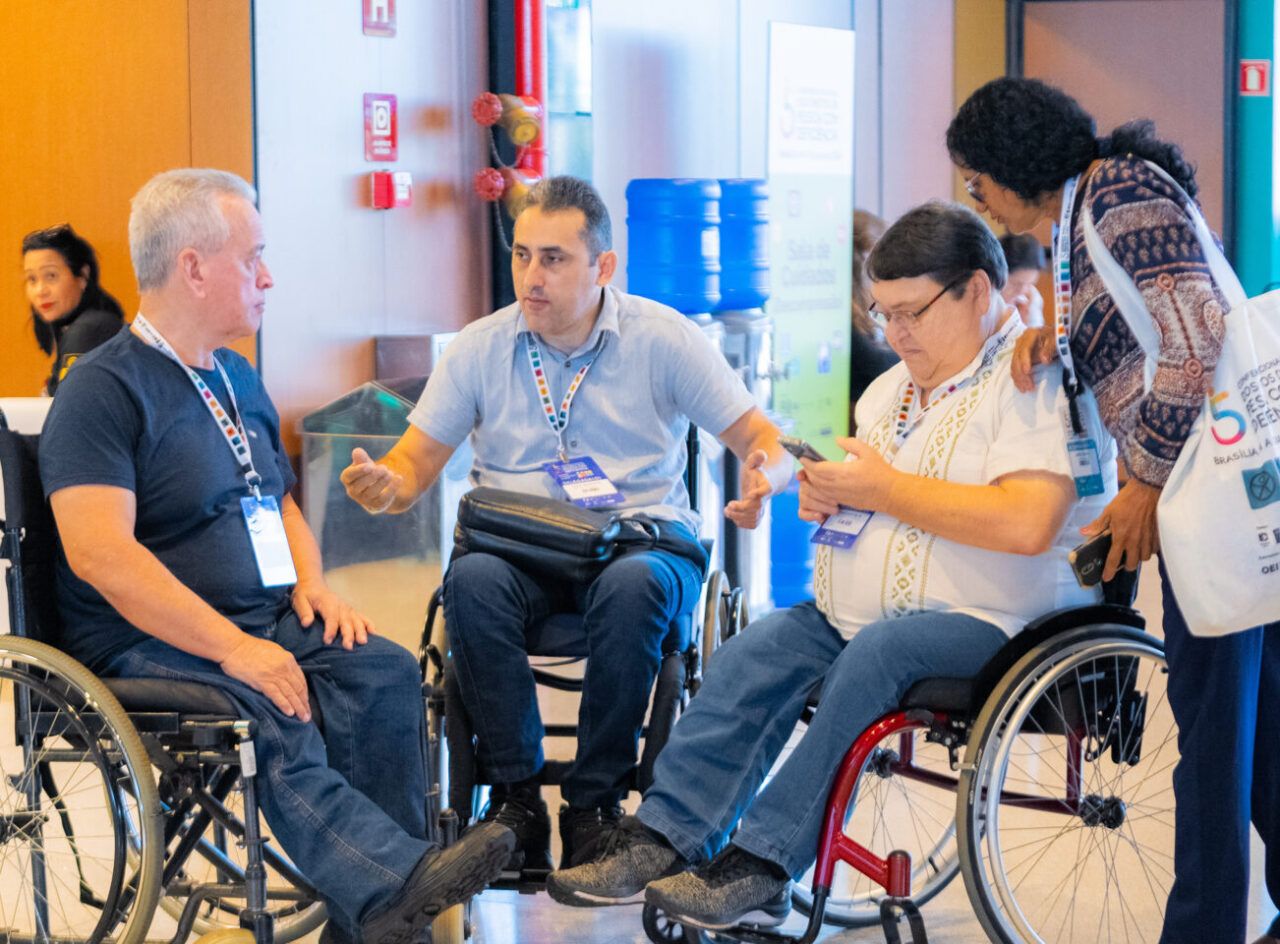 Três homens em cadeiras de rodas conversam em espaço interno de evento, enquanto uma mulher em pé interage com o grupo. Todos usam crachás e participam de atividade institucional.
