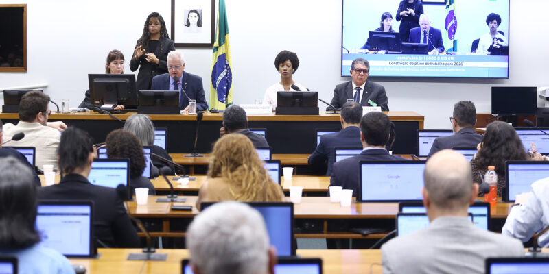 Sala de audiência com mesa principal ao fundo, onde autoridades participam de reunião institucional. Um homem fala ao microfone enquanto outras pessoas estão sentadas ao lado. Atrás, uma intérprete de Libras realiza a tradução. Ao fundo, há a bandeira do Brasil e um telão exibindo a transmissão da sessão. Em primeiro plano, participantes acompanham a atividade sentados, utilizando computadores portáteis.