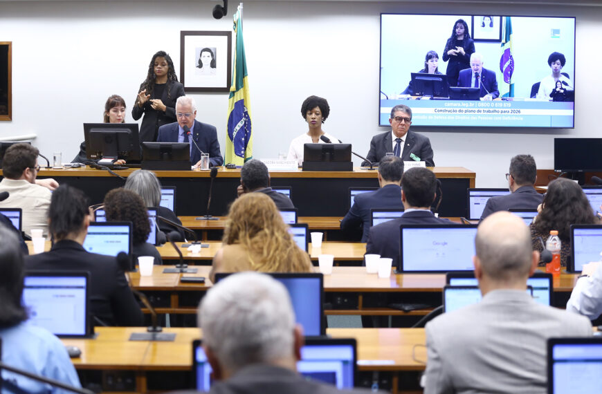 Sala de audiência com mesa principal ao fundo, onde autoridades participam de reunião institucional. Um homem fala ao microfone enquanto outras pessoas estão sentadas ao lado. Atrás, uma intérprete de Libras realiza a tradução. Ao fundo, há a bandeira do Brasil e um telão exibindo a transmissão da sessão. Em primeiro plano, participantes acompanham a atividade sentados, utilizando computadores portáteis.