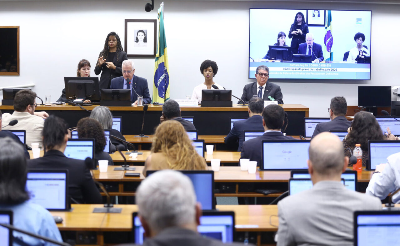 Sala de audiência com mesa principal ao fundo, onde autoridades participam de reunião institucional. Um homem fala ao microfone enquanto outras pessoas estão sentadas ao lado. Atrás, uma intérprete de Libras realiza a tradução. Ao fundo, há a bandeira do Brasil e um telão exibindo a transmissão da sessão. Em primeiro plano, participantes acompanham a atividade sentados, utilizando computadores portáteis.