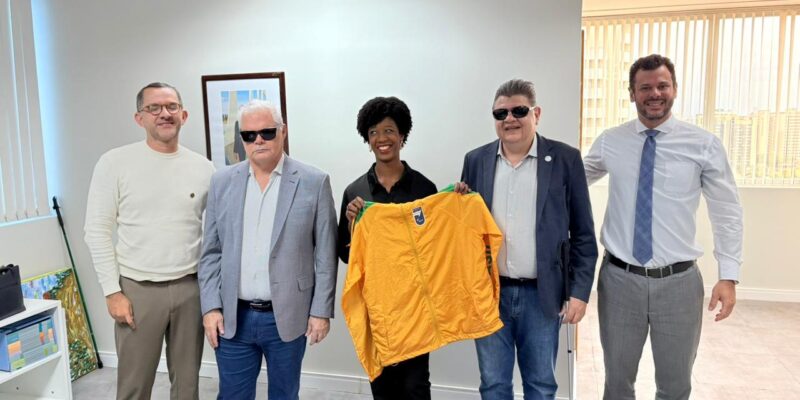 A secretária nacional Isadora Nascimento posa ao lado de representantes do Comitê Paralímpico Brasileiro (CPB) em sala de reuniões, enquanto segura uma jaqueta esportiva amarela ao centro do grupo, com móveis e paredes claras ao fundo.