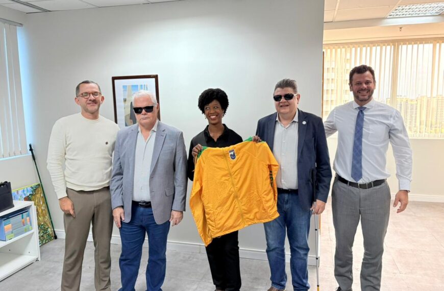 A secretária nacional Isadora Nascimento posa ao lado de representantes do Comitê Paralímpico Brasileiro (CPB) em sala de reuniões, enquanto segura uma jaqueta esportiva amarela ao centro do grupo, com móveis e paredes claras ao fundo.