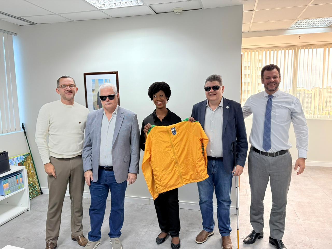 A secretária nacional Isadora Nascimento posa ao lado de representantes do Comitê Paralímpico Brasileiro (CPB) em sala de reuniões, enquanto segura uma jaqueta esportiva amarela ao centro do grupo, com móveis e paredes claras ao fundo.