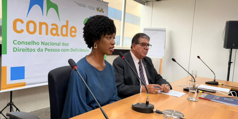 Isadora Nascimento, mulher negra, fala ao microfone em reunião, usando blusa azul e brincos dourados. Ao lado, Roberto Paulo do Vale Tiné, de terno e óculos. Ao fundo, banner do Conade.