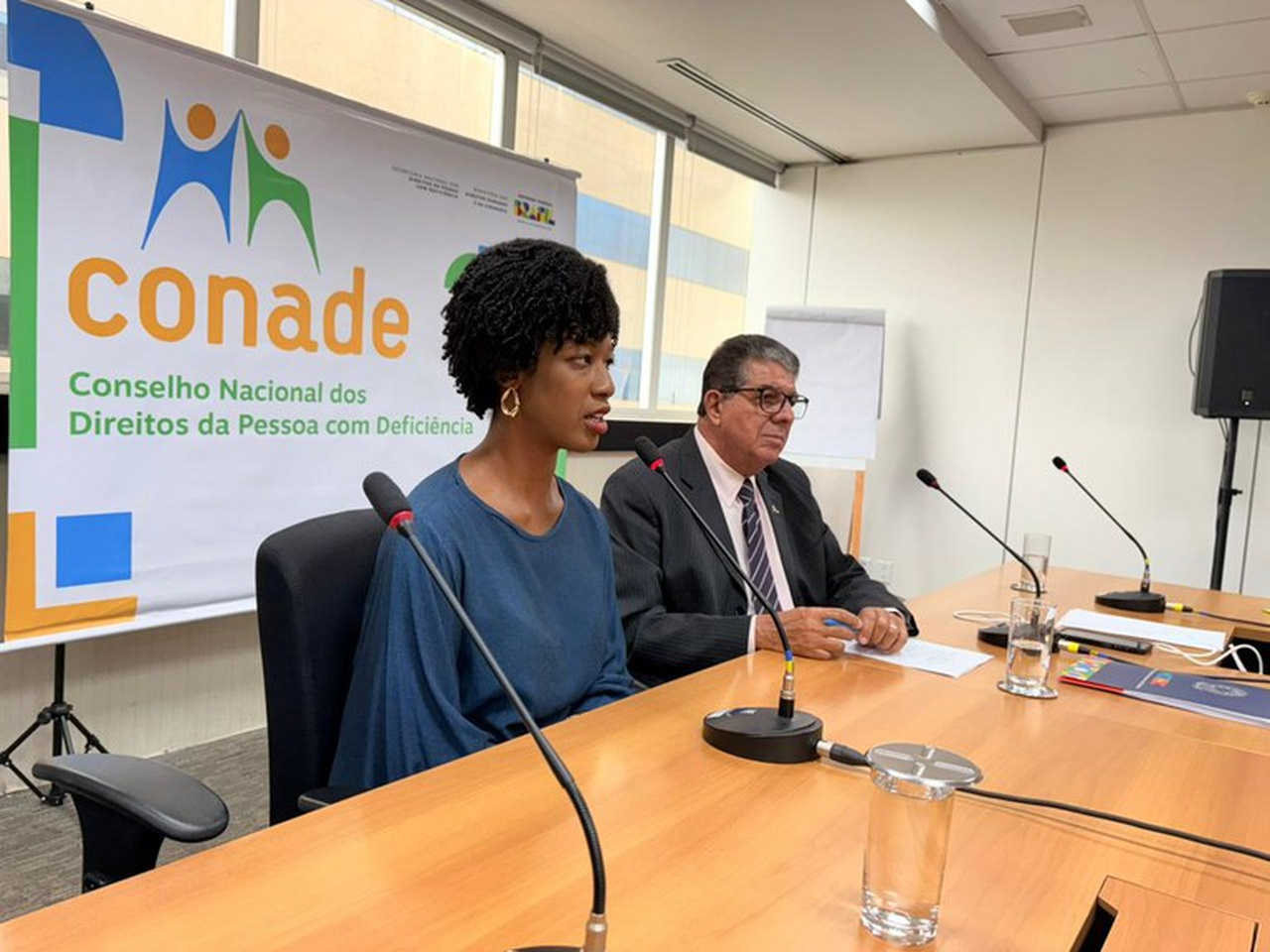 Isadora Nascimento, mulher negra, fala ao microfone em reunião, usando blusa azul e brincos dourados. Ao lado, Roberto Paulo do Vale Tiné, de terno e óculos. Ao fundo, banner do Conade.