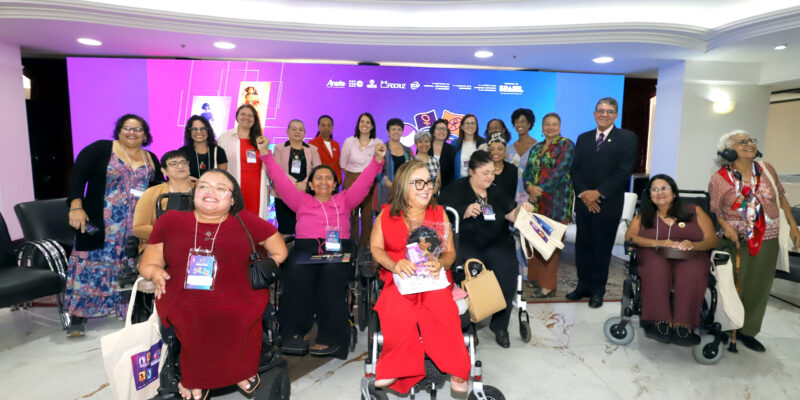 Grupo de mulheres com e sem deficiência posa em evento institucional. Em destaque, participantes em cadeiras de rodas na primeira fila. Ao fundo, painel colorido com logotipos oficiais.