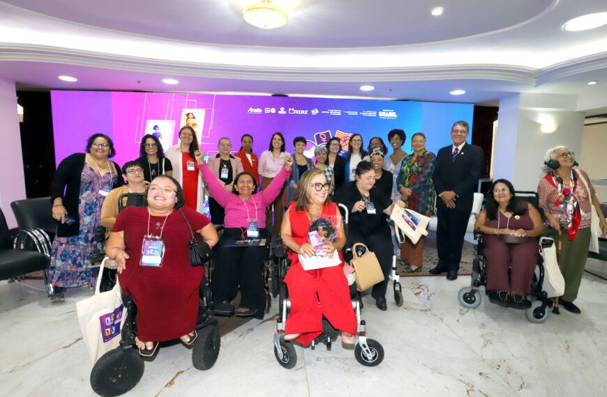 Grupo de mulheres com e sem deficiência posa em evento institucional. Em destaque, participantes em cadeiras de rodas na primeira fila. Ao fundo, painel colorido com logotipos oficiais.