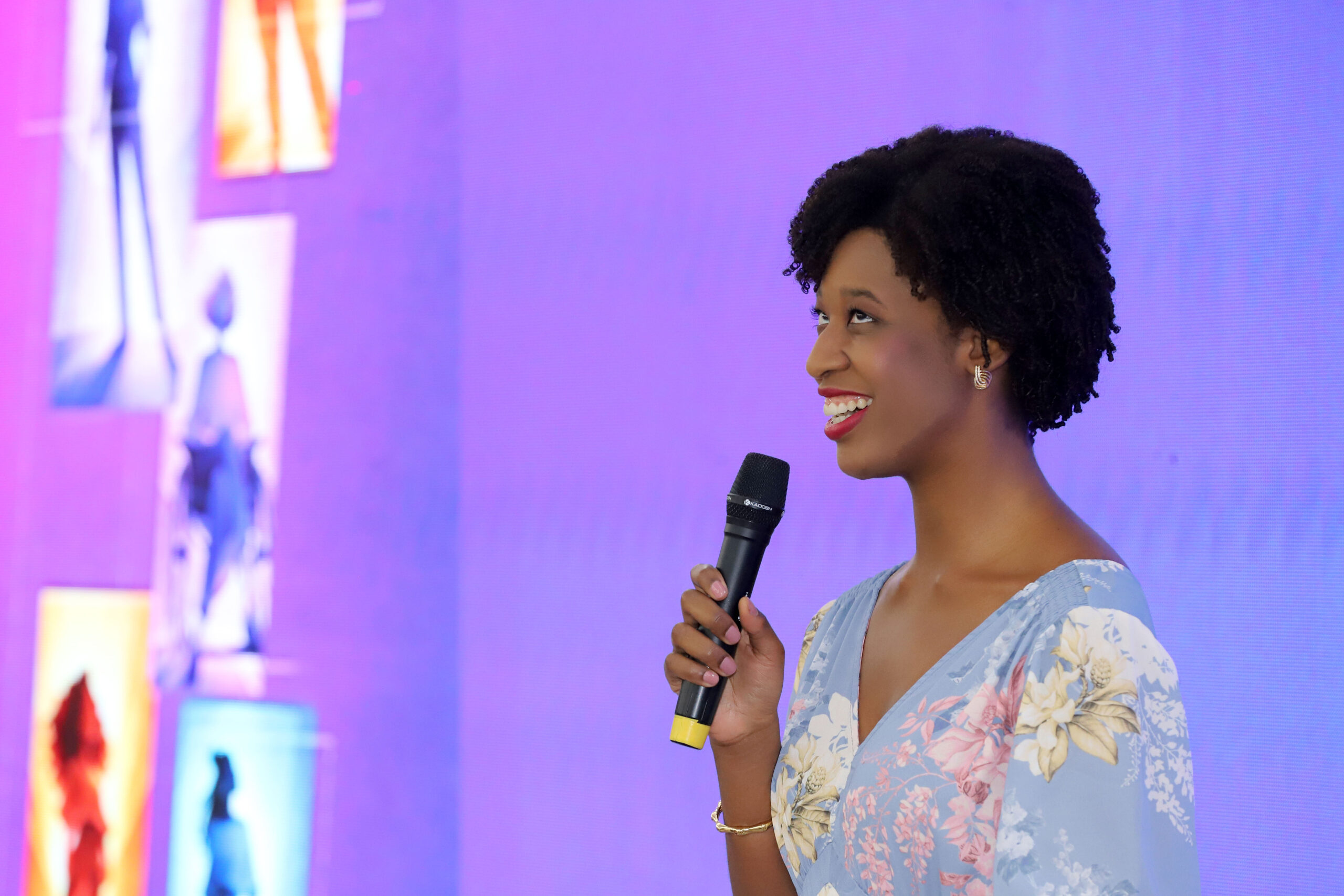 Encontro Nacional fortalece protagonismo de mulheres com deficiência e impulsiona agenda de inclusão no país Mulher negra, de cabelo curto crespo, segura microfone e sorri durante fala. Usa vestido floral azul claro. Ao fundo, painel em tons de roxo e rosa com imagens estilizadas.